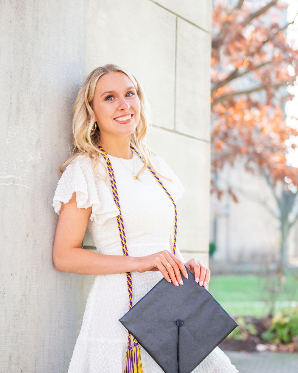 A graduate in white dress poses with their diploma cap against a light wall with autumn foliage in background.