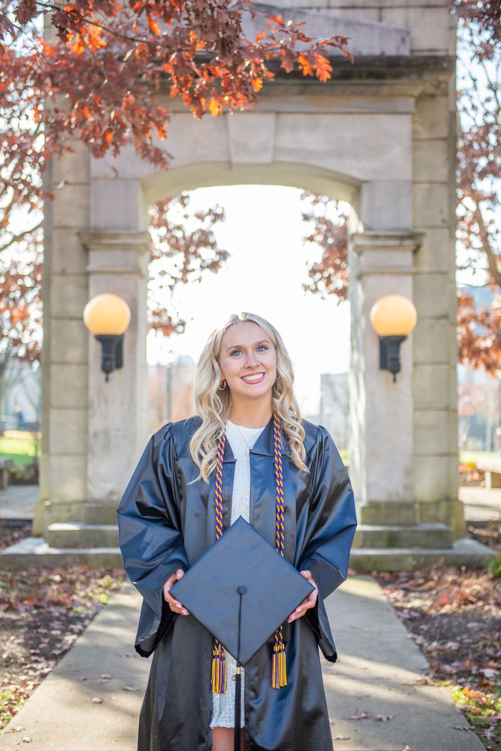 Graduate in blue cap and gown poses in front of stone archway with autumn leaves during graduation photos.
