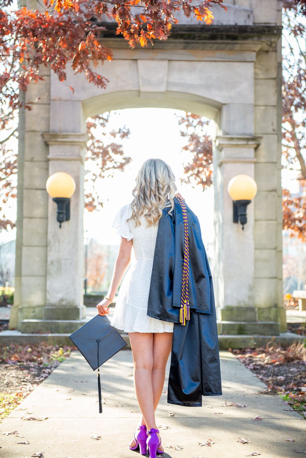Graduate in blue gown and white dress holds cap in front of stone arch with glowing lamps and autumn leaves.