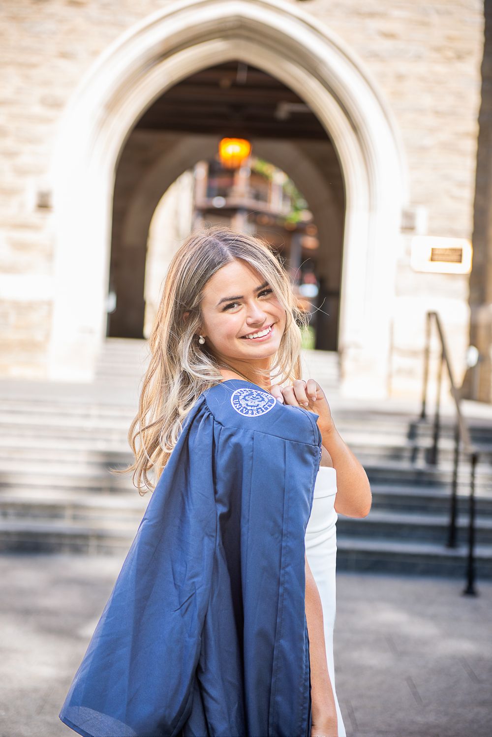 Graduation photo featuring a graduate holding up a blue academic gown while standing under a stone archway.
