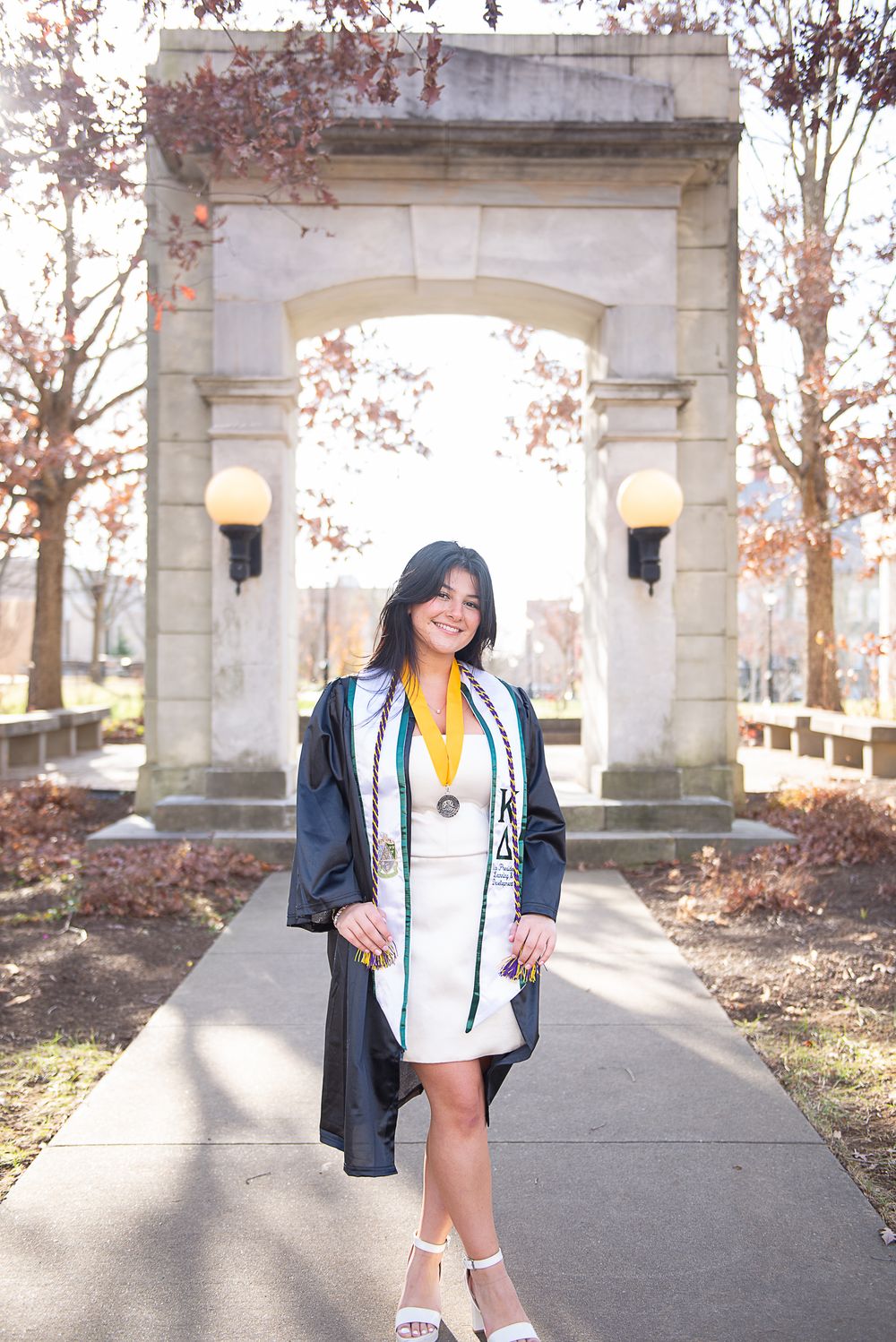 Graduate poses in decorated cap and gown at stone archway during golden hour lighting.