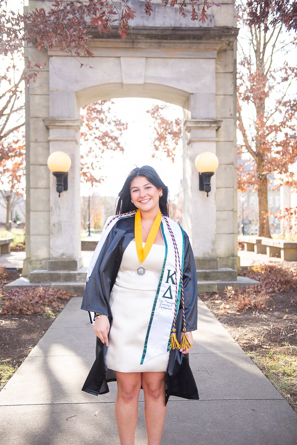 Graduate in white dress holding academic sash and medal poses under stone archway during autumn season.