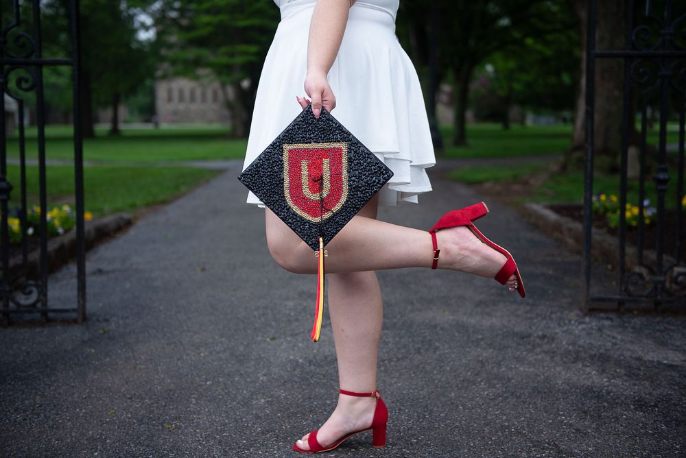 Graduate in white dress lifts leg playfully on campus path while holding Indiana University graduation sign and wearing red heels.
