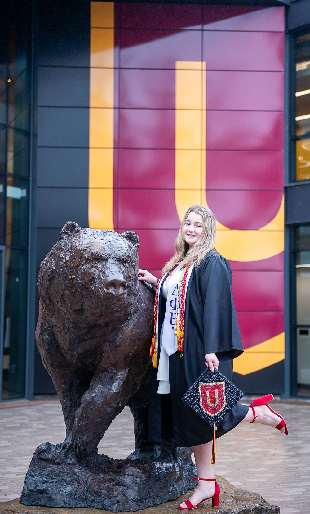 Graduate poses with bronze bear statue in front of a red and gold wall while holding a graduation cap.