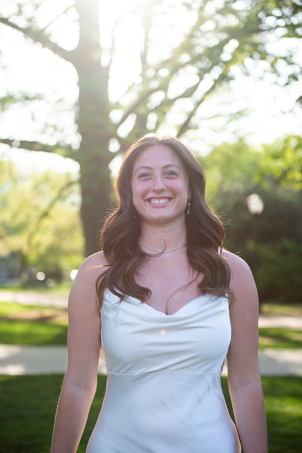 Person in white sleeveless dress smiles warmly while standing outdoors on sunny day with trees in background.