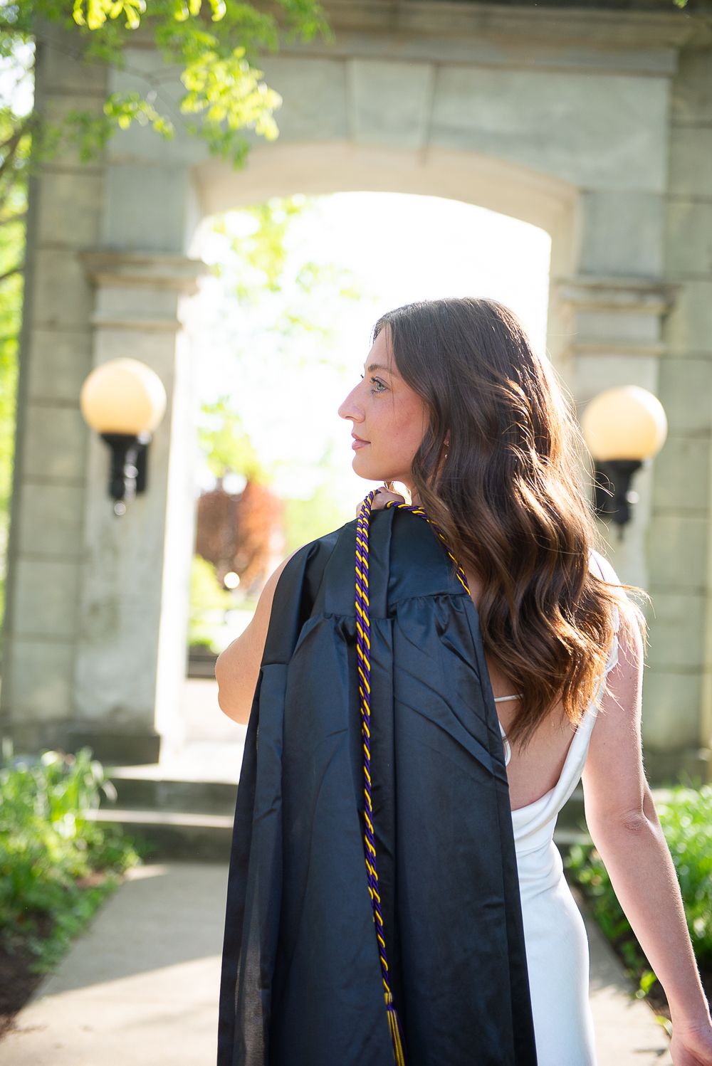 Graduate in cap and gown stands in front of stone archway with glowing sunset light and vintage lamps.