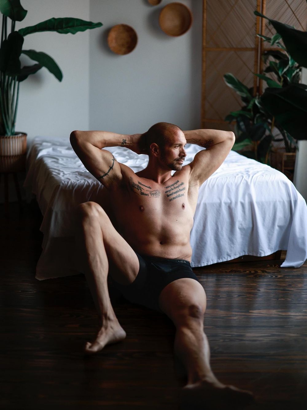 Contemporary artistic nude photography against a dark wooden floor with natural light and houseplants in the background.