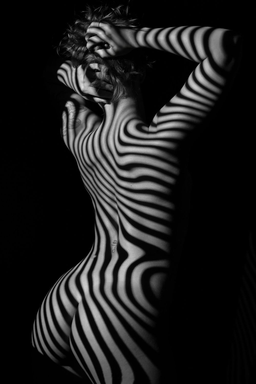 Artistic nude with projected zebra-like striped shadows creating an abstract black and white pattern effect.