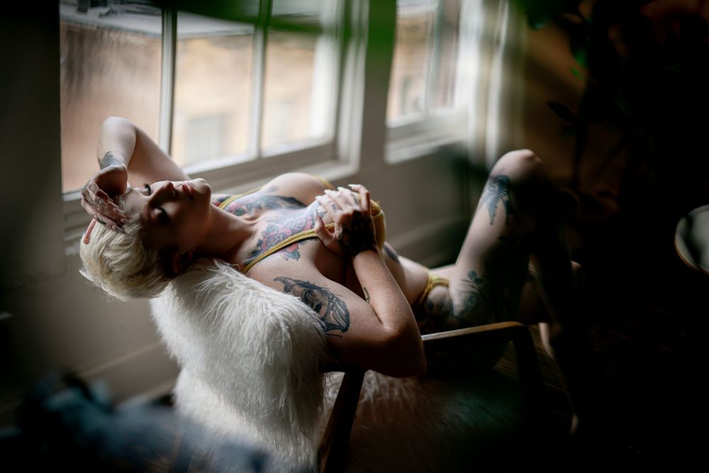 Tattooed woman reclining in a chair by a sunlit window in artistic boudoir photography.