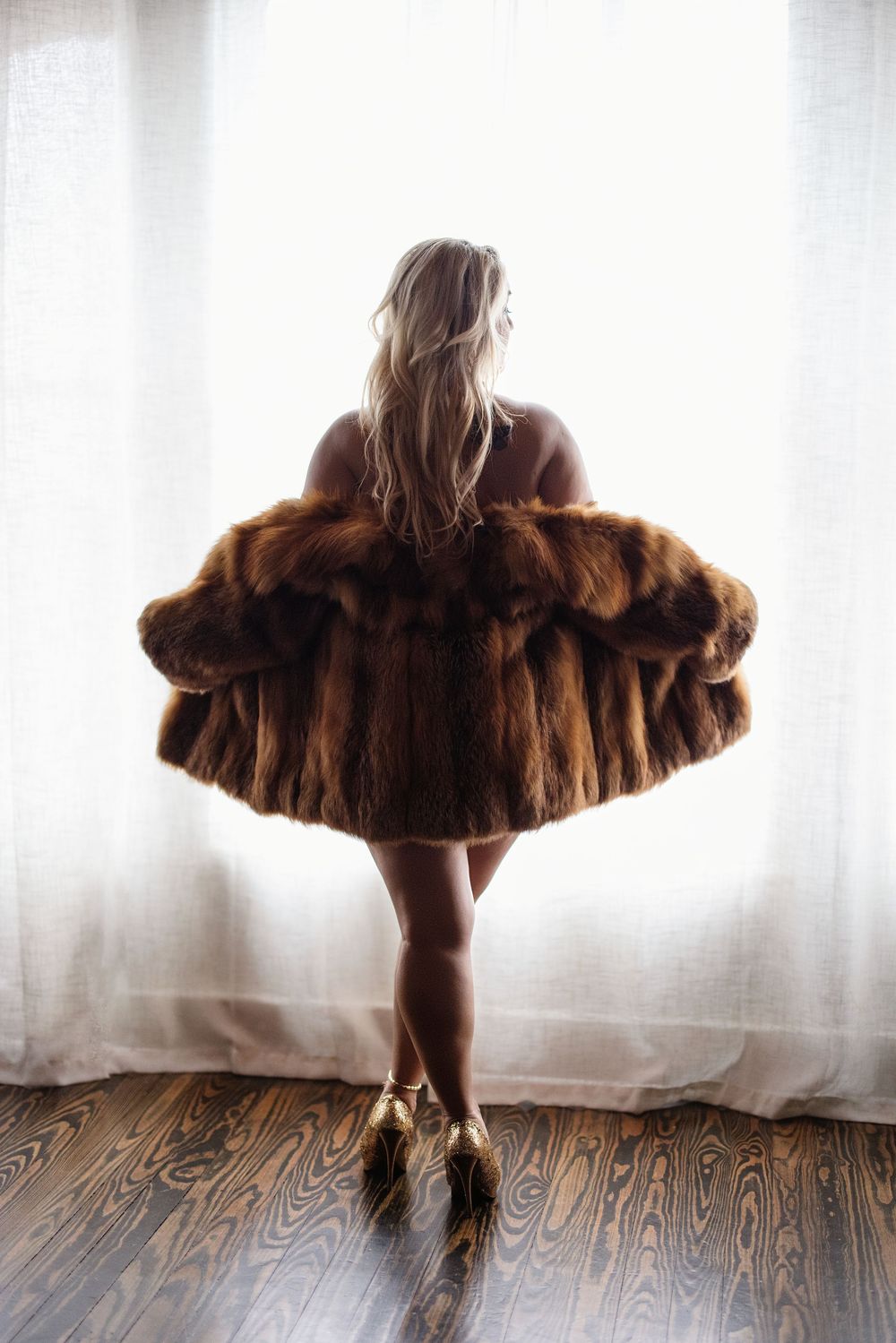 Woman in luxurious brown fur coat standing by sheer curtains on hardwood floor, viewed from behind.