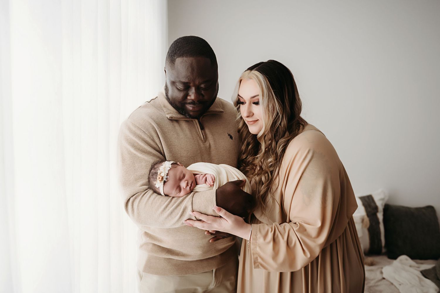 A couple in matching beige outfits holding and lovingly gazing at their newborn baby at home.
