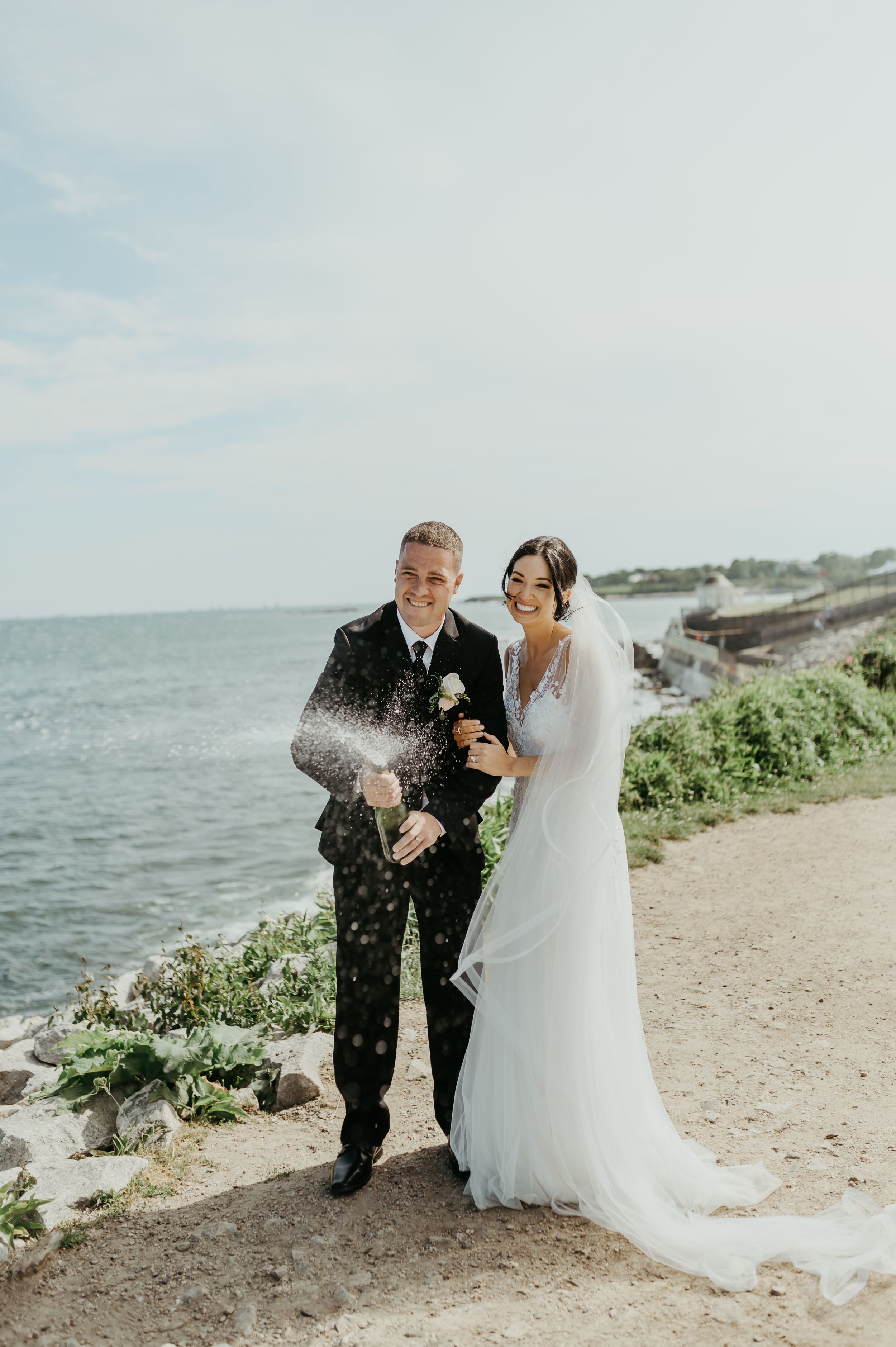 A couple celebrates their wedding day by popping champagne on a rocky coastal shoreline.