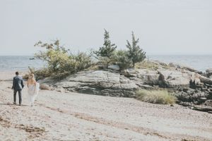 Newlyweds walk hand in hand along a rocky beach shoreline with coastal vegetation.