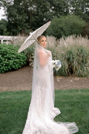 A bride in a lace wedding gown holds a white parasol while standing in a garden with ornamental grasses. Farmington Polo Club, CT
