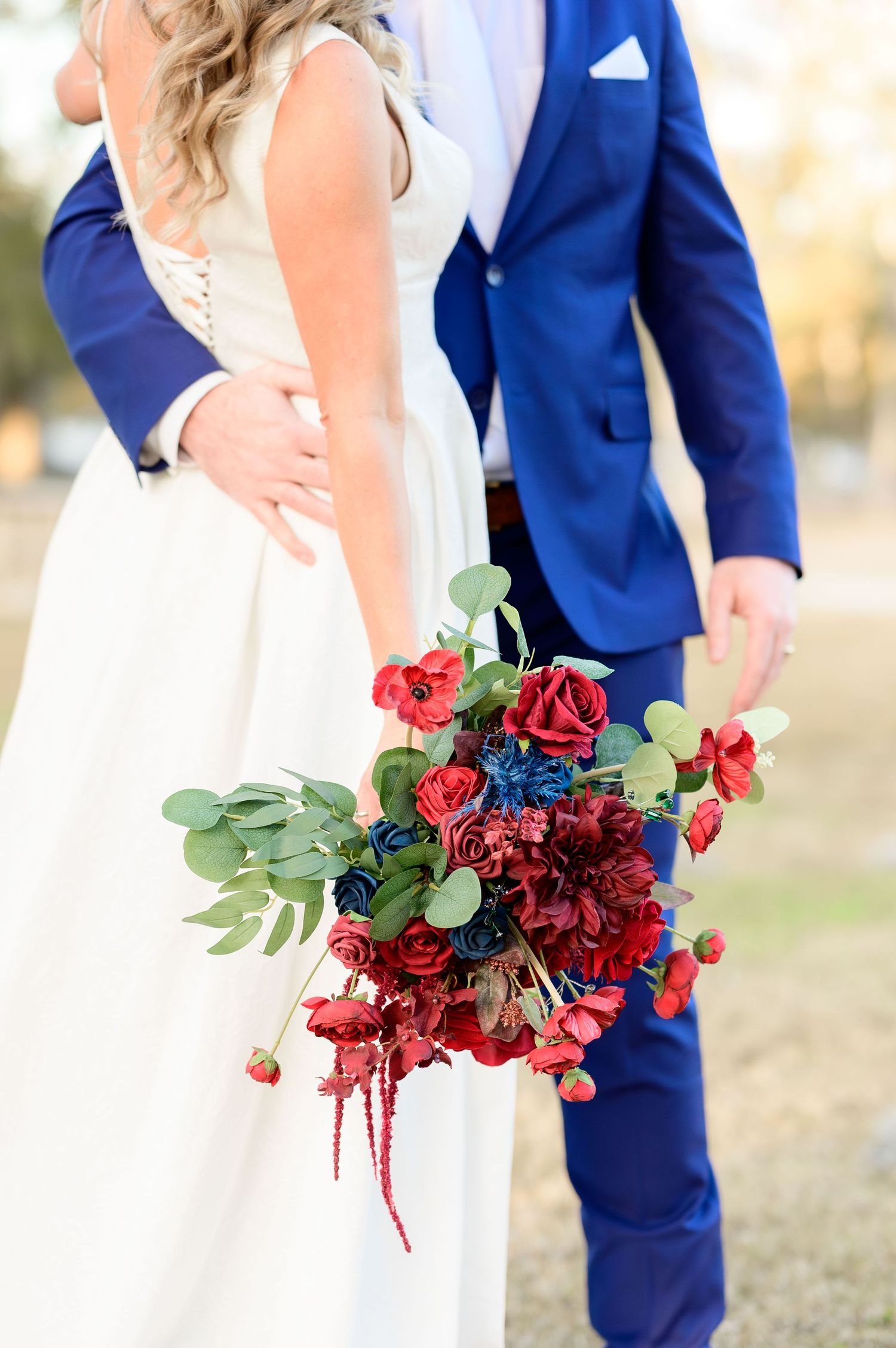 Close-up of bridal bouquet featuring red roses and eucalyptus against blue suit and white dress.