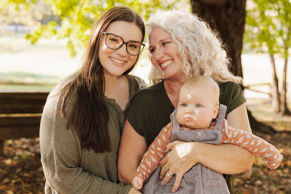 Three generations share a happy outdoor moment together in warm sunlight under trees.