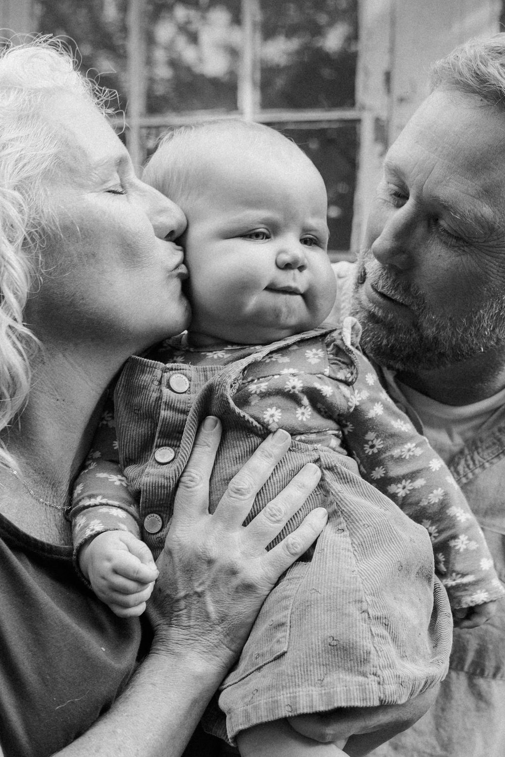 Black and white photo showing loving grandparents kissing their chubby-cheeked granddaughter in corduroy overalls.