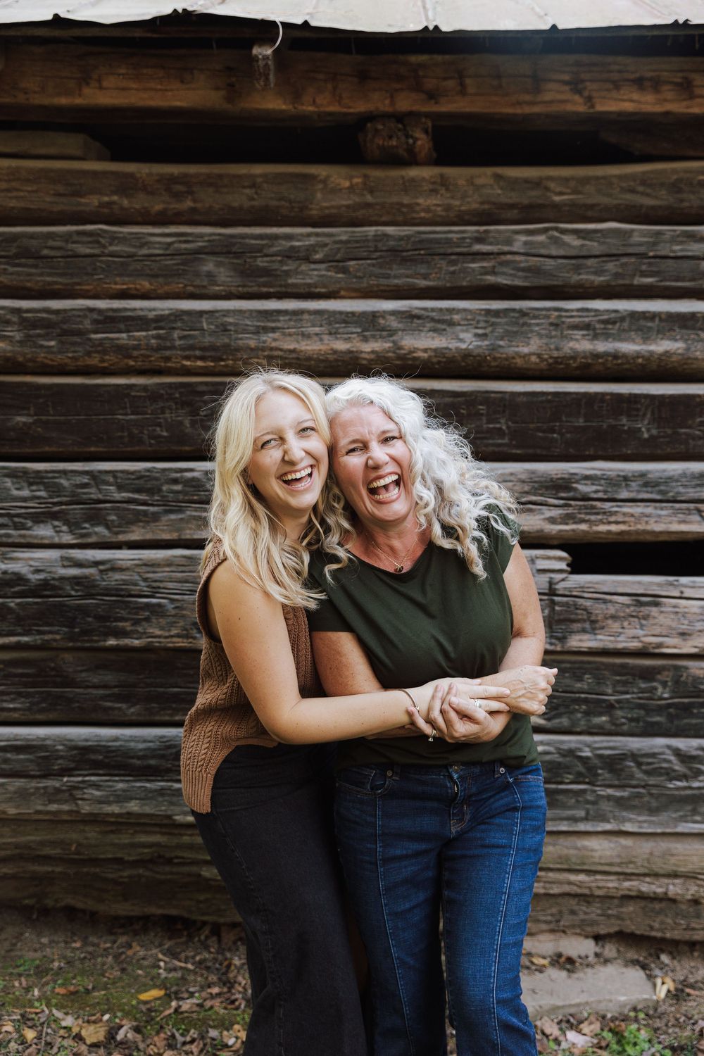 Two people laughing together joyfully against a rustic wooden log cabin wall.