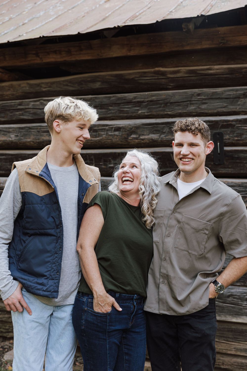Mom and her two sons share a candid laughing moment while posing together against a rustic wooden wall.