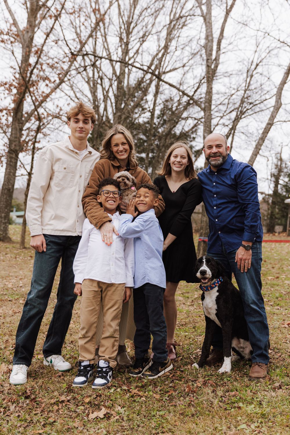 A family stands together outdoors in autumn with their black and white dog among bare trees and fallen leaves.