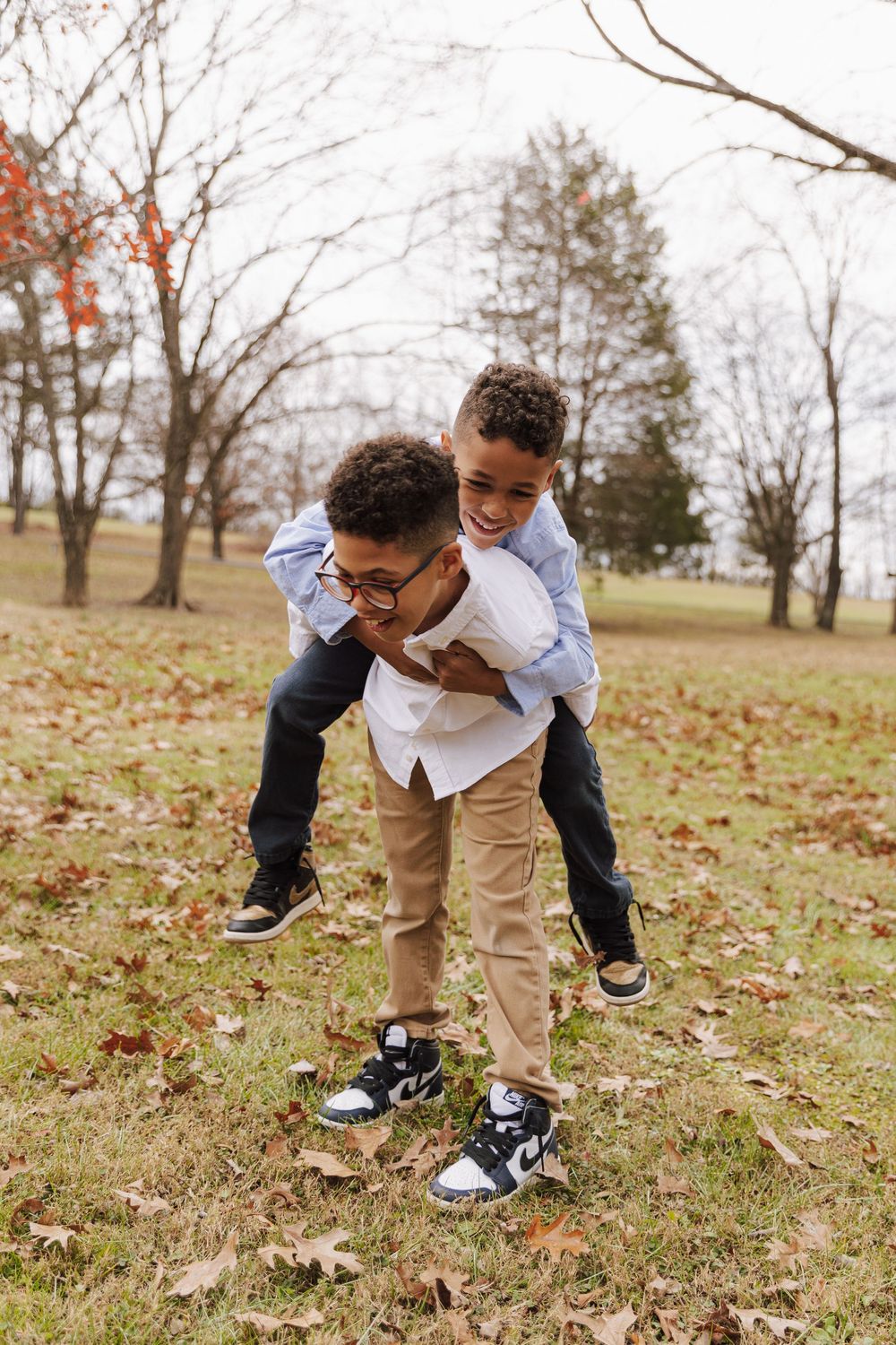 Two young boys playing and piggybacking on a fall day in a park with fallen leaves on the ground.