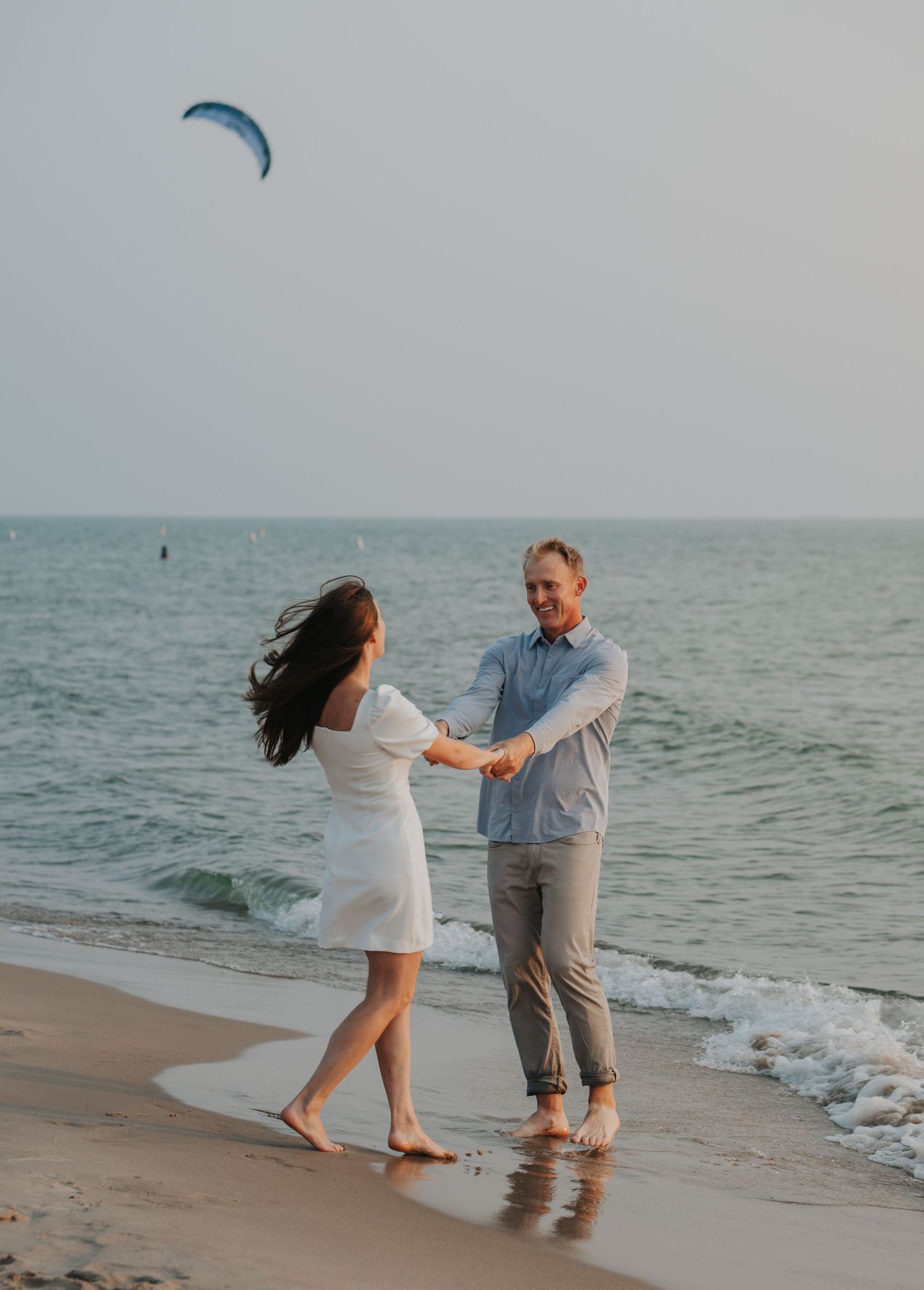 A couple dances barefoot on a sandy beach shore, with a kite soaring in the sky above the ocean.