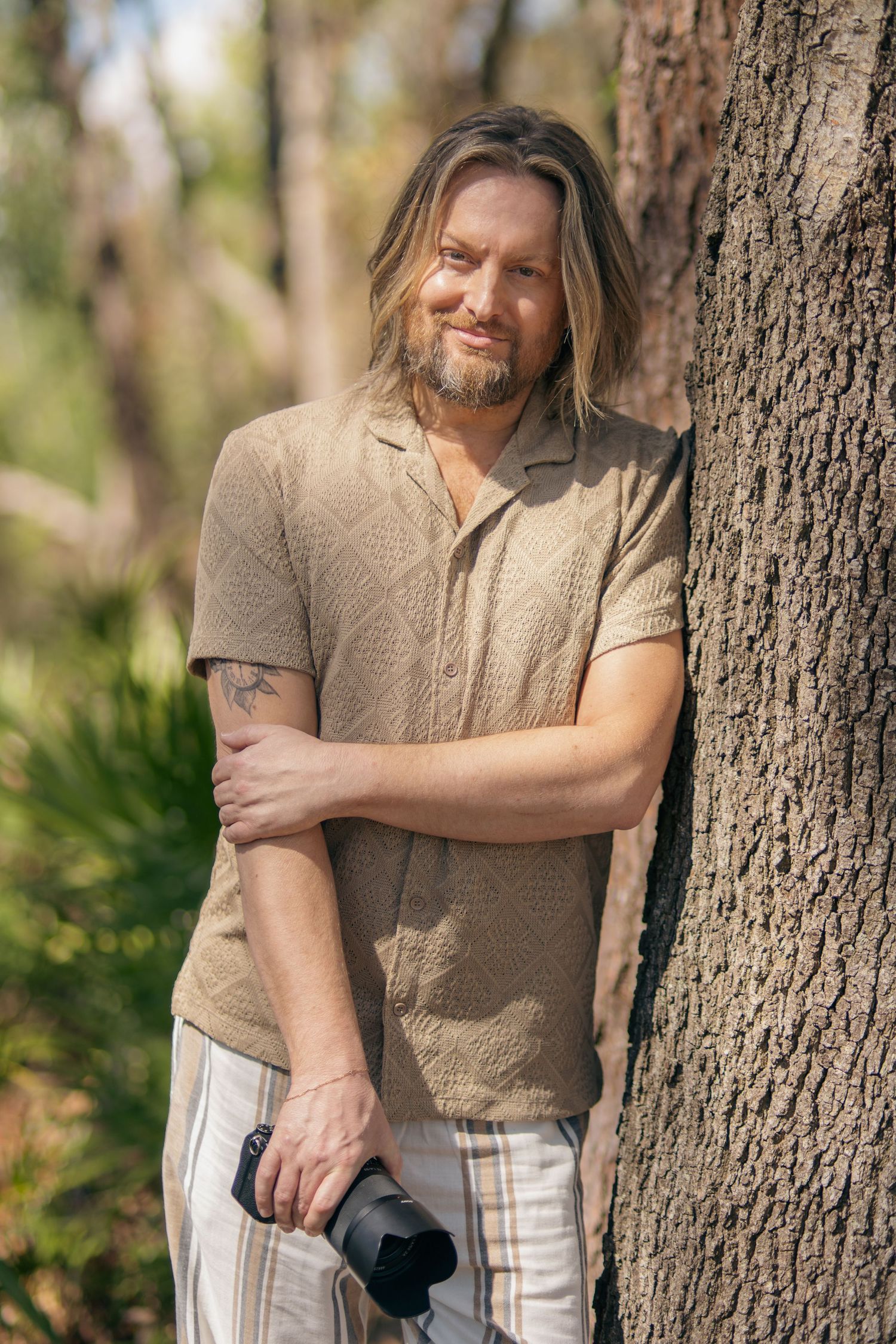 A person in casual attire leaning against a tree trunk outdoors.