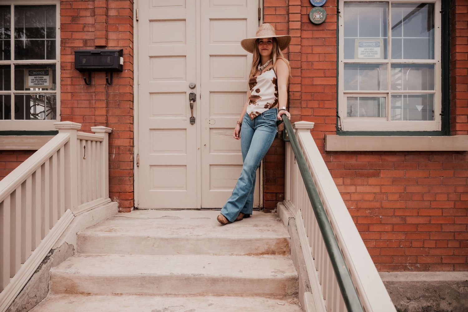 Summer western fashion shoot featuring a model in a brown cow print silk top and straw hat against a brick background.