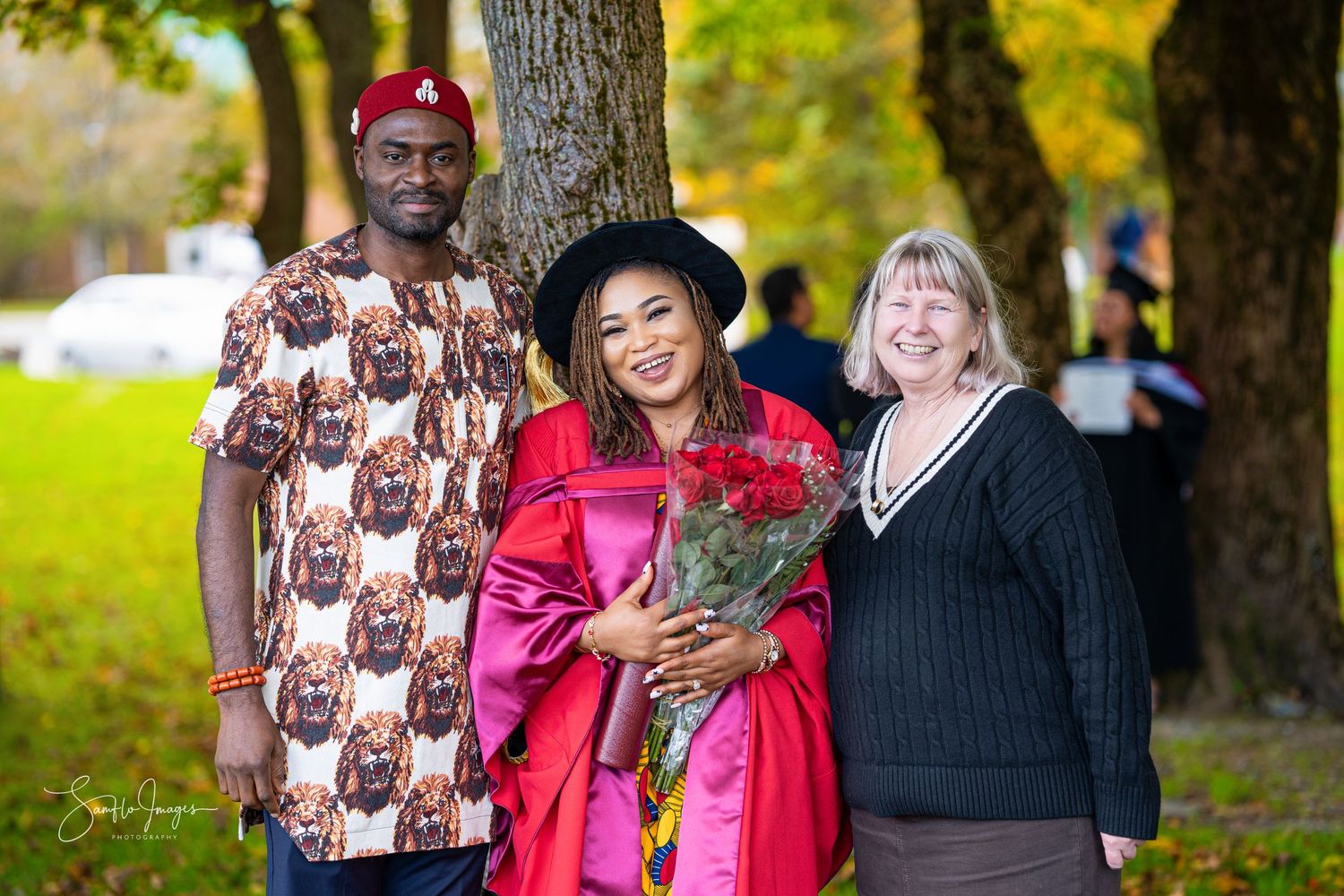 Three people celebrating a graduation in red academic regalia with flowers pose outdoors under autumn trees.