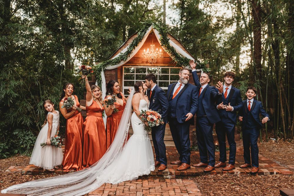 A wedding party poses in front of a rustic wooden chapel with bridesmaids in rust-colored dresses.