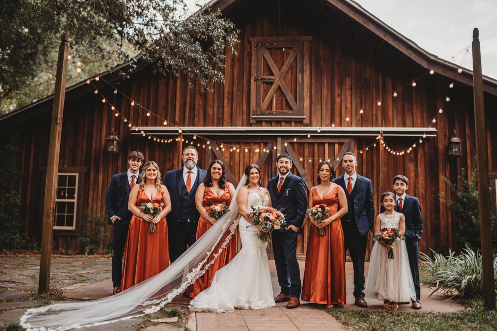 Wedding party group photo in front of a rustic barn with string lights and burnt orange bridesmaid dresses.