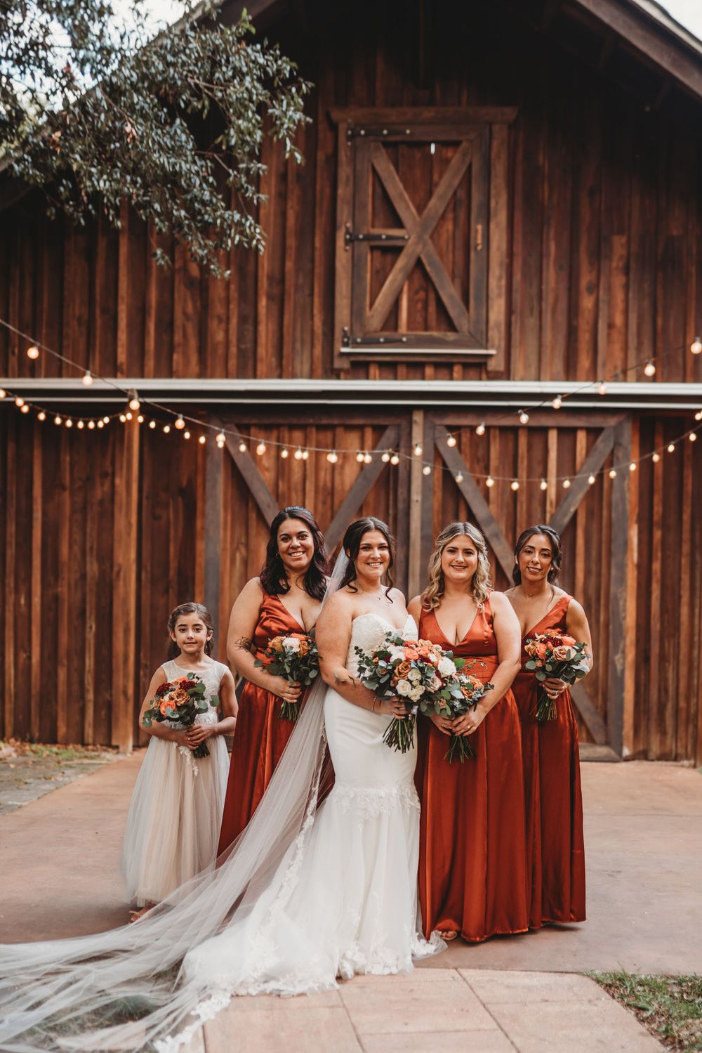 Group photo of bridesmaids in rust-colored dresses with the bride in front of a wooden barn.