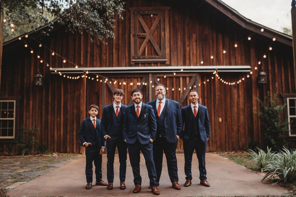 Groomsmen in navy suits pose in front of a rustic barn with string lights.