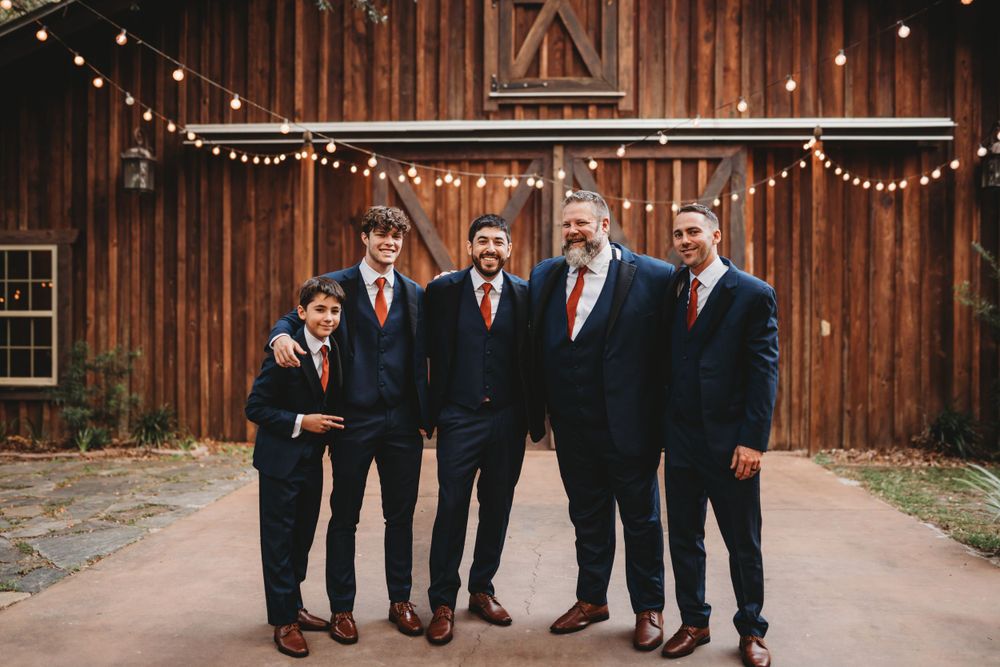 Groomsmen in dark suits with red ties pose together in front of a rustic wooden barn.
