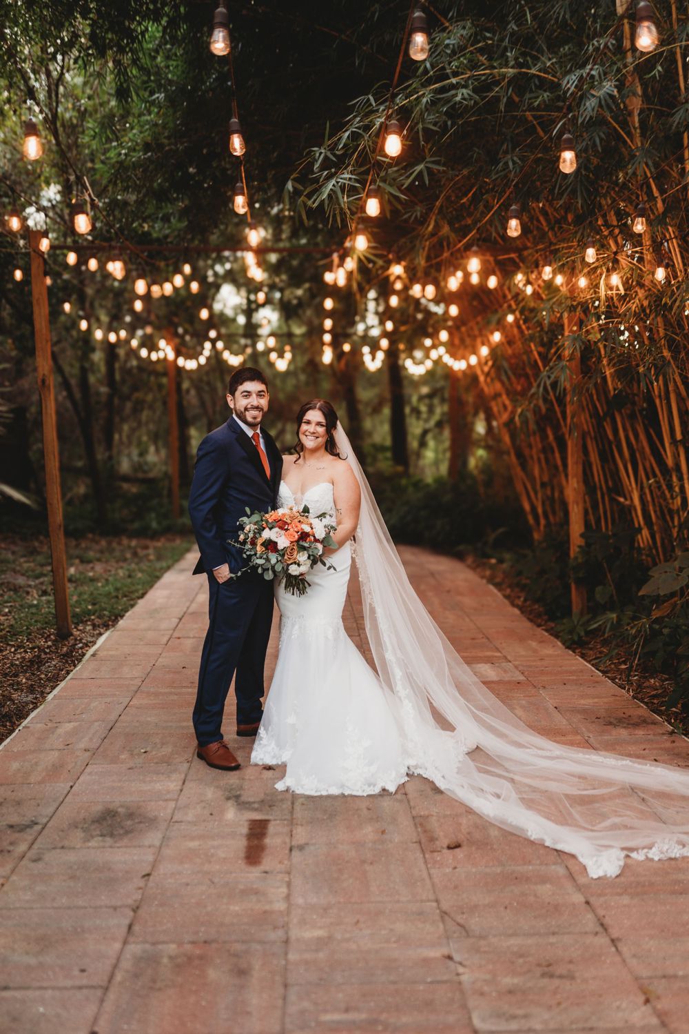 A romantic evening portrait of newlyweds under string lights on a brick pathway.