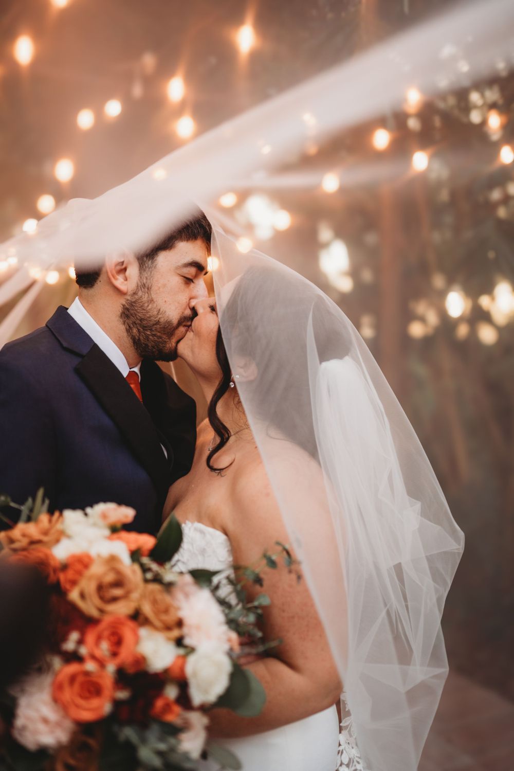 A romantic wedding kiss under twinkling lights with the bride holding a coral and white rose bouquet.