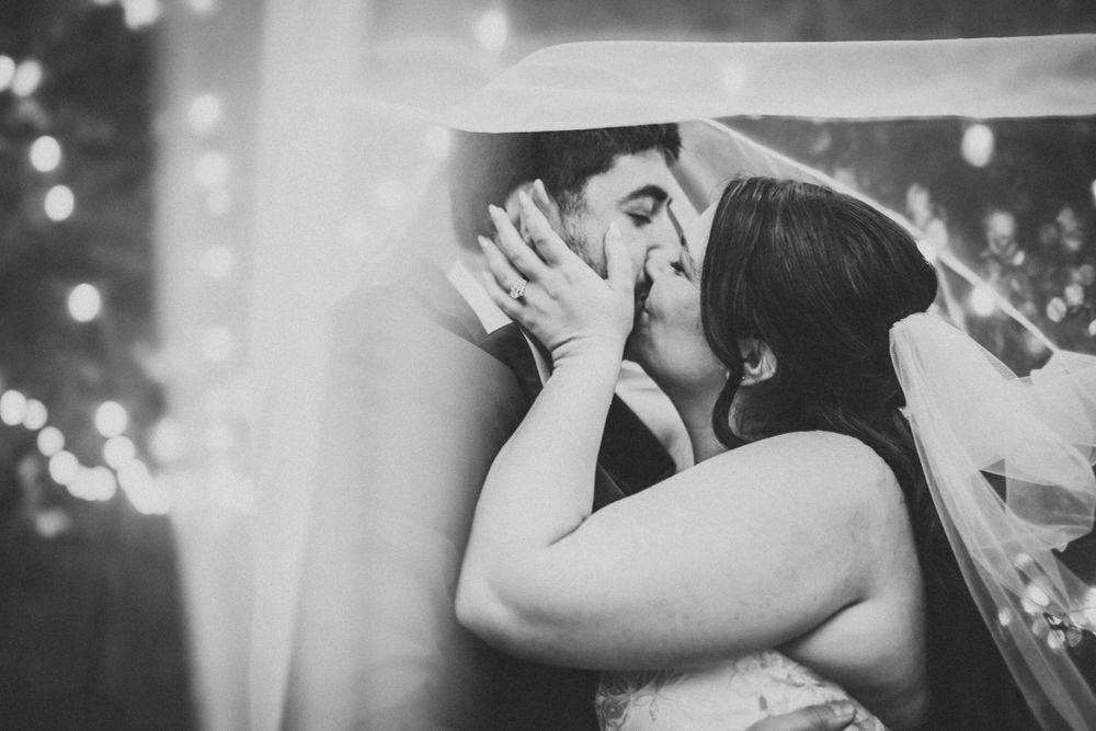 Intimate black and white photo of a wedding couple sharing a tender kiss with string lights in background.