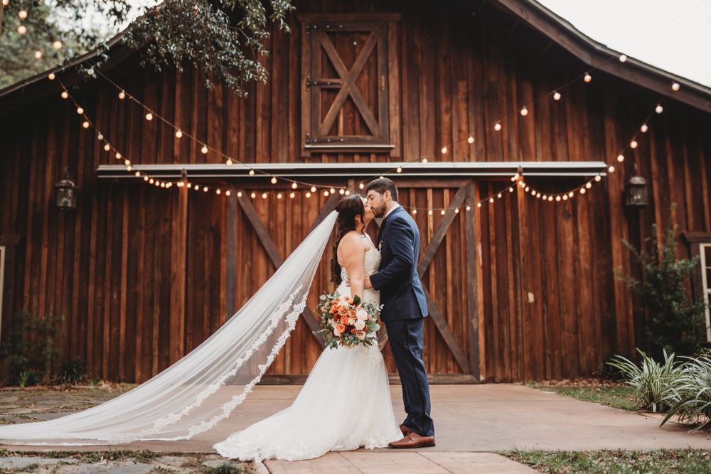Newlywed couple kisses in front of a rustic barn decorated with string lights.
