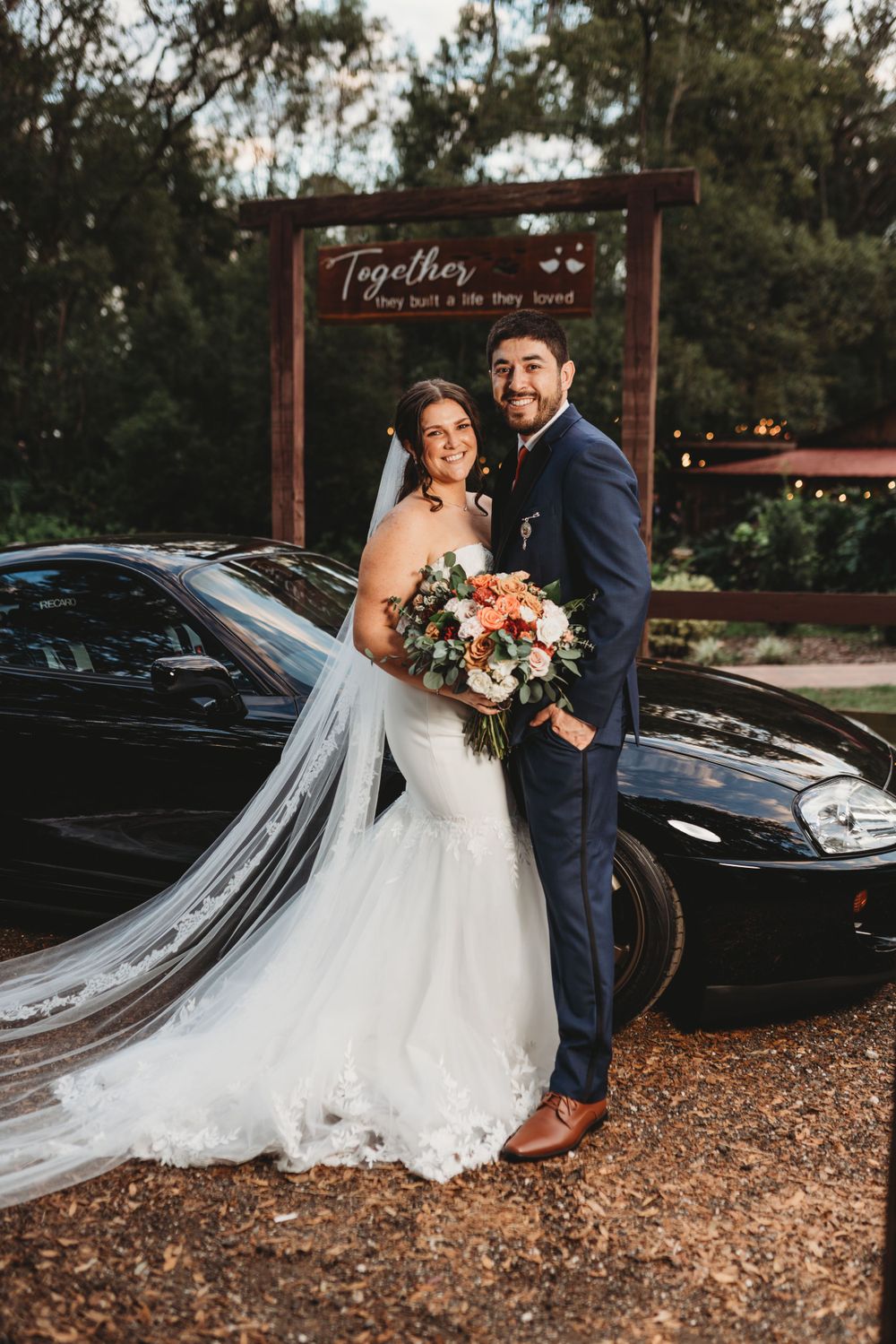 Couple poses in wedding attire in front of a wooden arch with 'Together' sign.