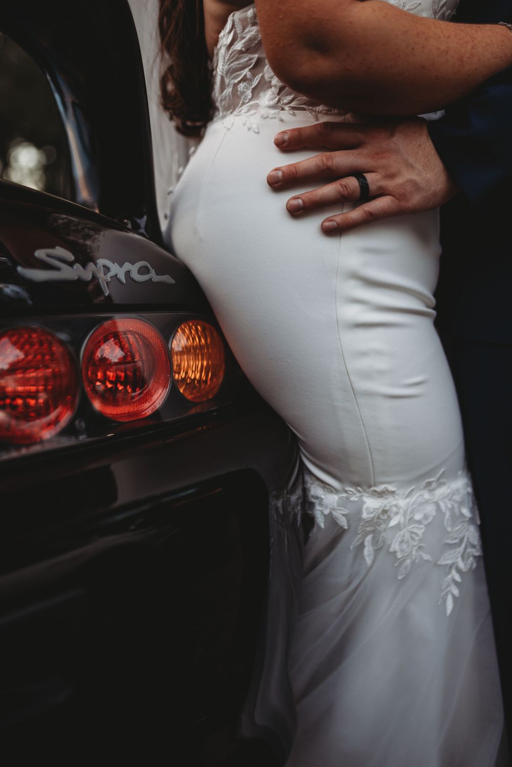 Close-up of a bride's fitted white wedding dress with lace details near car taillights.