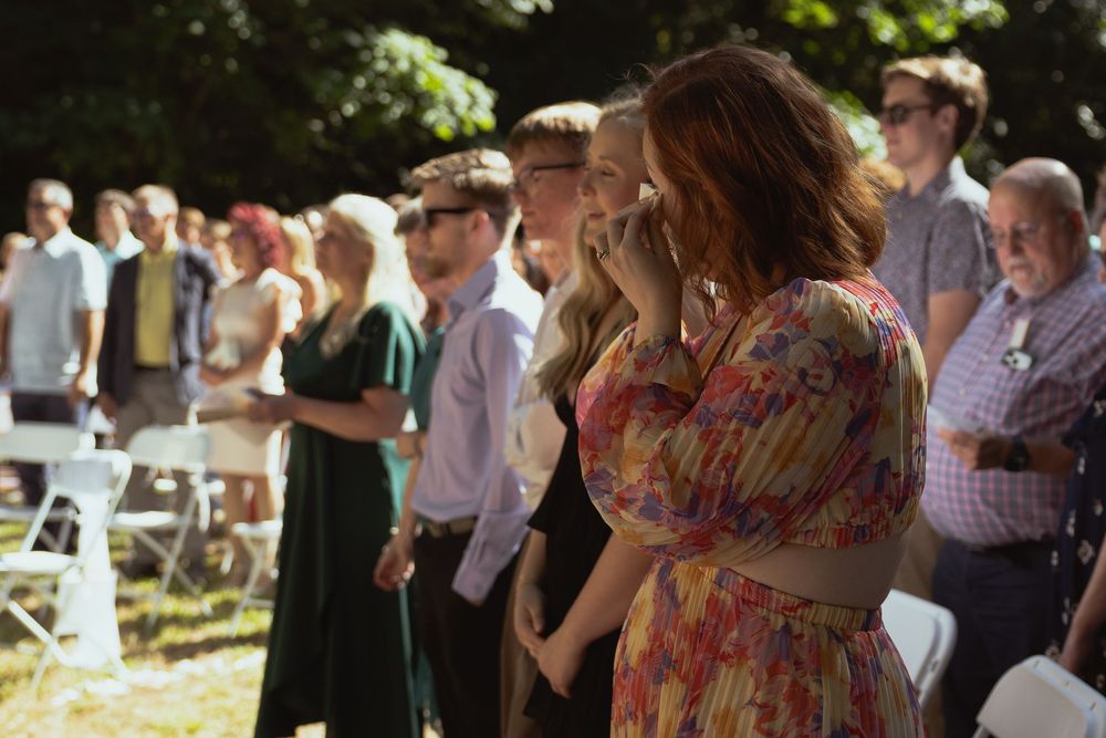 Emotional wedding guest in floral dress wiping tears during outdoor ceremony with crowd of attendees.