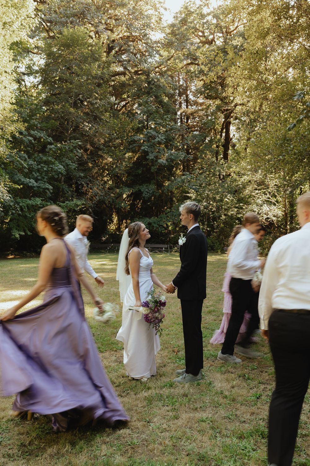 Bride and groom holding hands outdoors while wedding guests in purple dresses move around them in a sunlit garden.