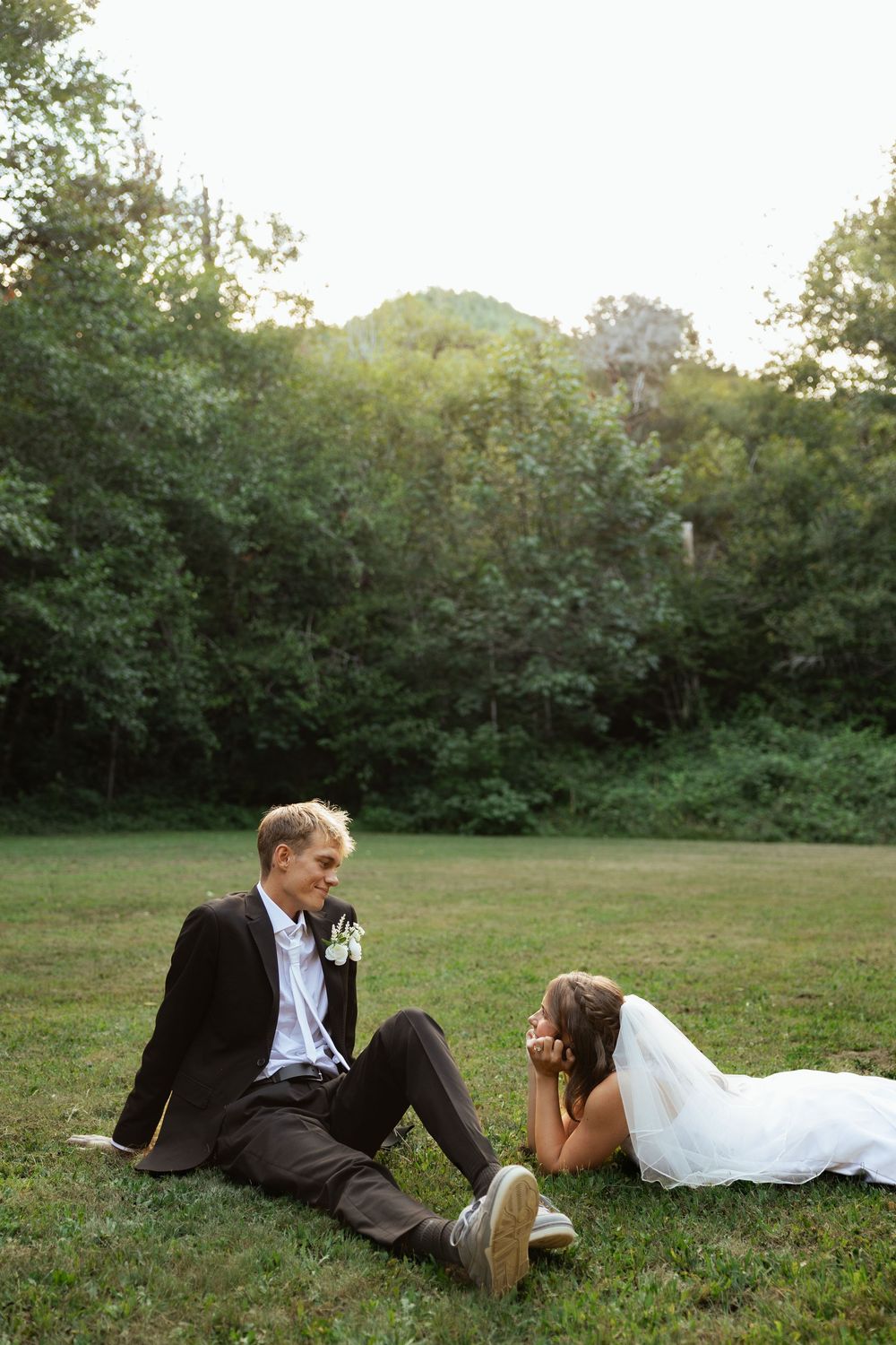 Bride and groom sitting on green grass in a lush outdoor setting, sharing a romantic moment in wedding attire.