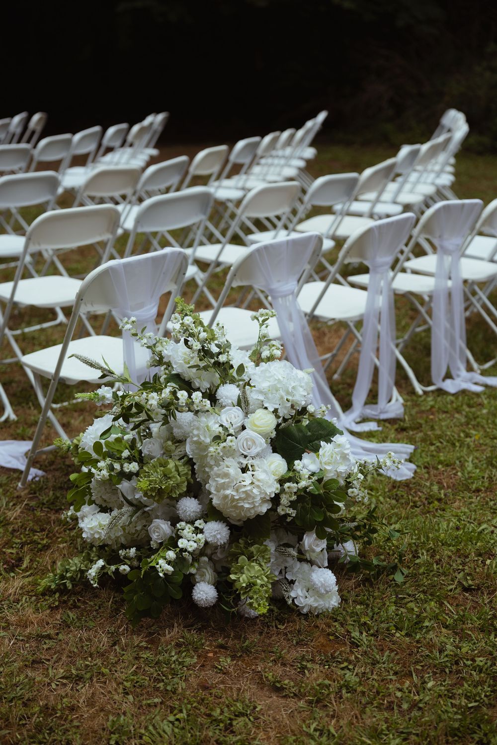 White folding chairs arranged in rows on grass for an outdoor wedding ceremony with white floral bouquet in foreground.