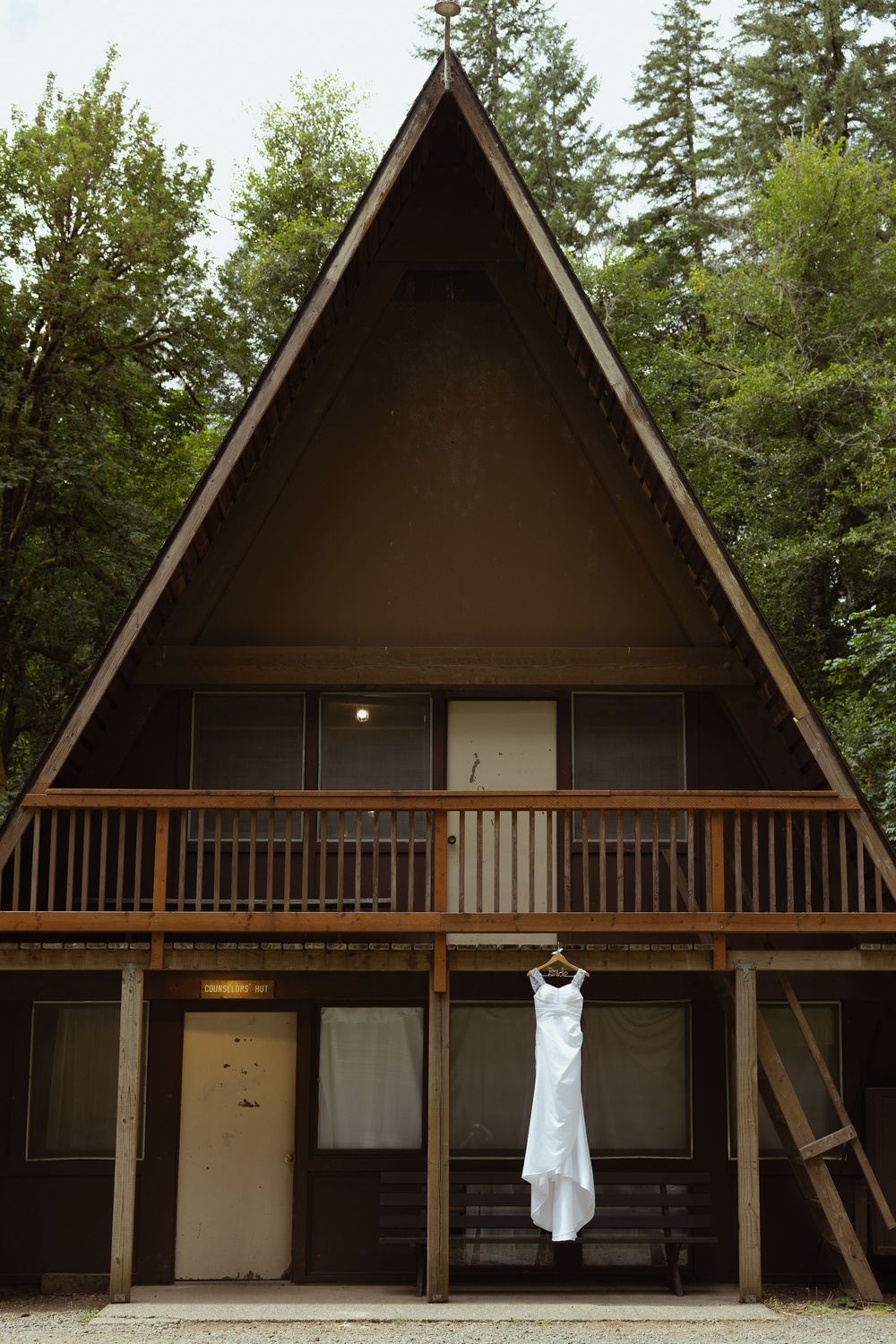 White wedding dress hanging on an A-frame cabin balcony surrounded by tall pine trees in a forest setting.