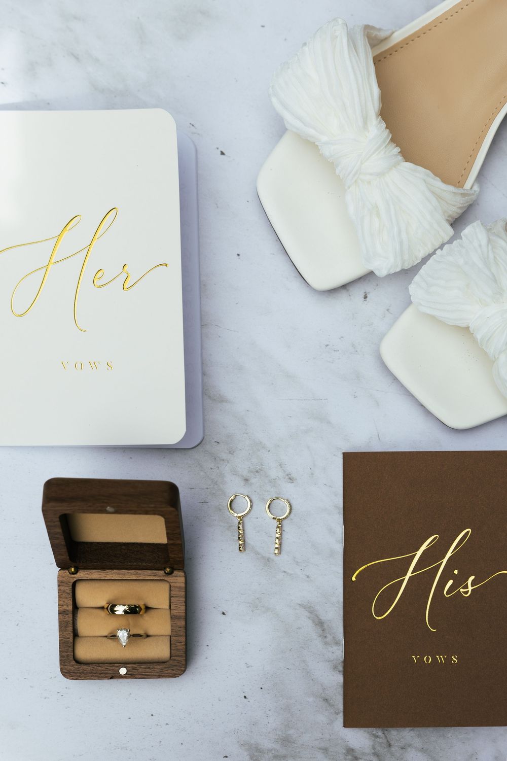Flat lay of wedding details including vow books, gold rings, earrings, and white bridal heels on marble.