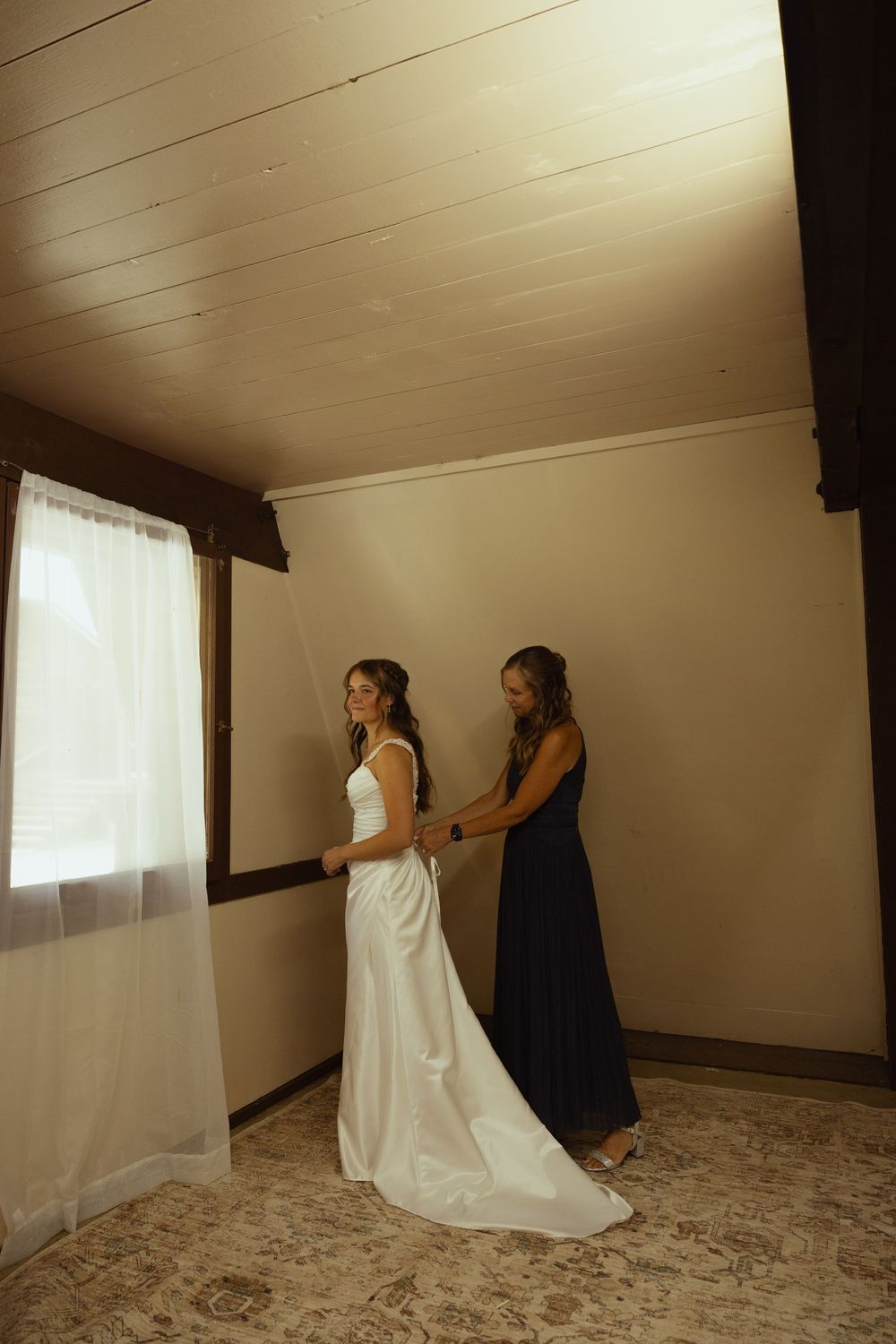 A bridesmaid in navy blue helps a bride in a white gown with her dress in a softly lit rustic room.