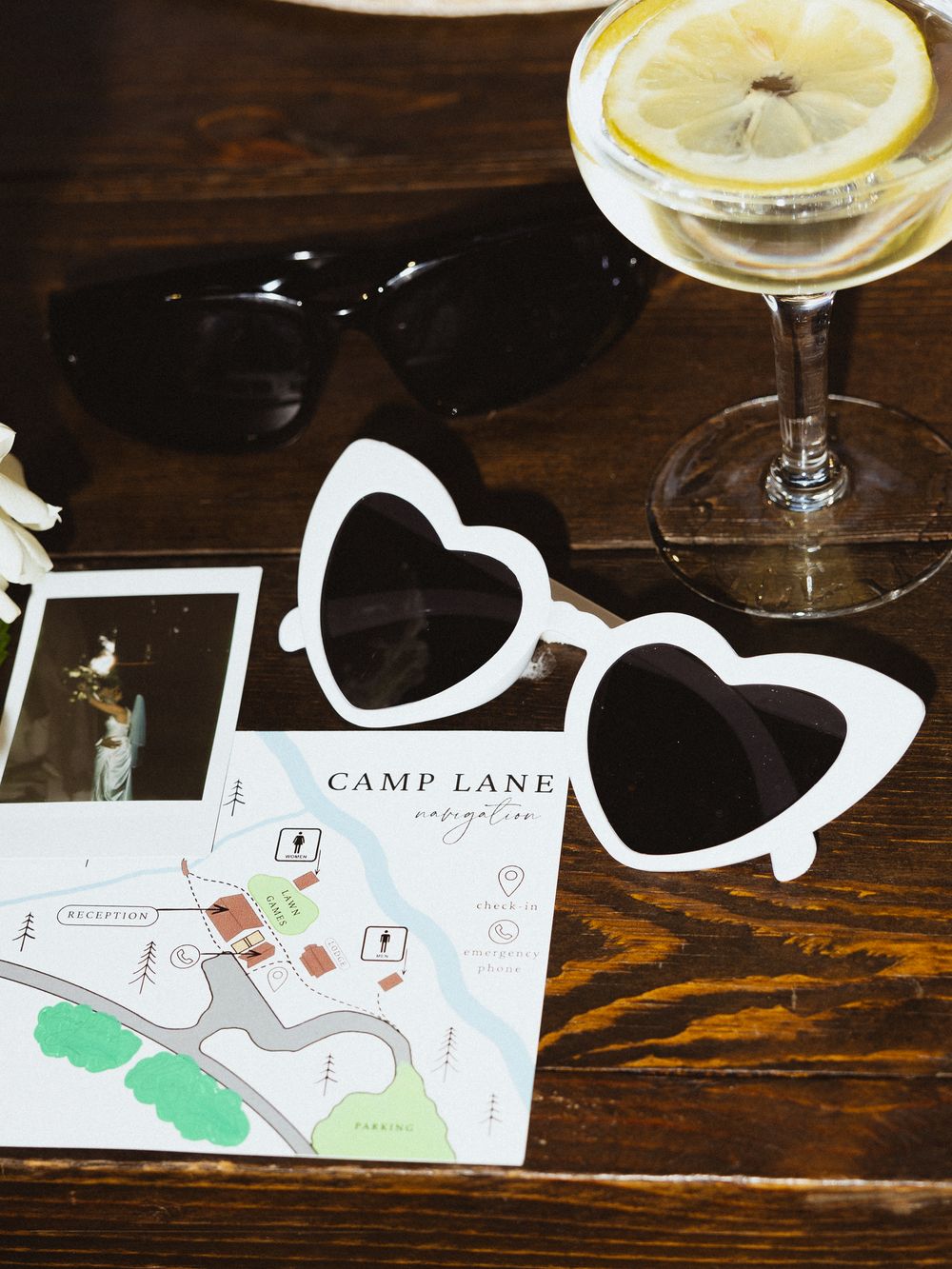 Heart-shaped white sunglasses and black sunglasses on a Camp Lane navigation map beside a lemon cocktail and Polaroid photos.