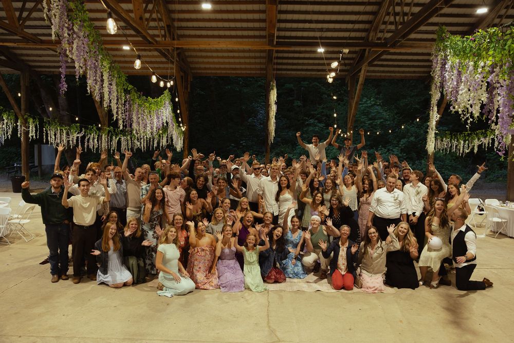 Large wedding group photo under floral decorations and string lights in an outdoor venue at night.