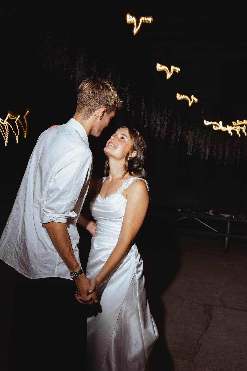Bride and groom holding hands and sharing a romantic moment at night with glowing fairy lights in the background.